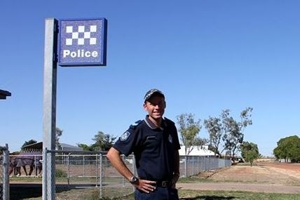 A police officer standing on front of a police sign in an remote outback town.