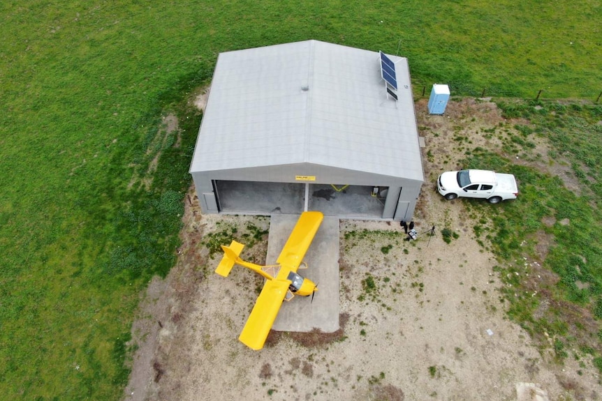 An aerial shot of a small yellow plan in front of a double-door hangar shed on a green paddock.