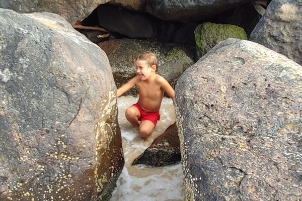 A boy with red shorts sitting between two rocks at the beach.