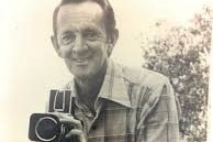 Black and white image of Bryan Hodgkinson stands behind a camera.