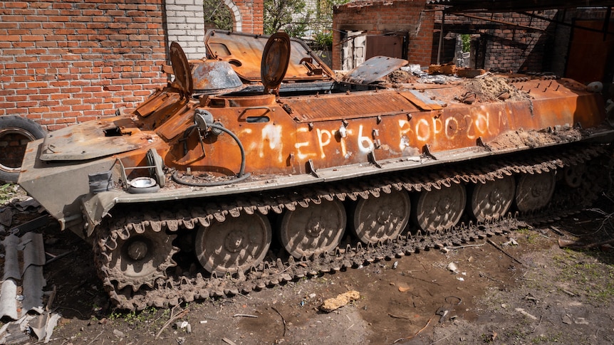 A tank stands stationary on the dirt next to brick buildings. It appears to be burnt out, with letters marked on the side