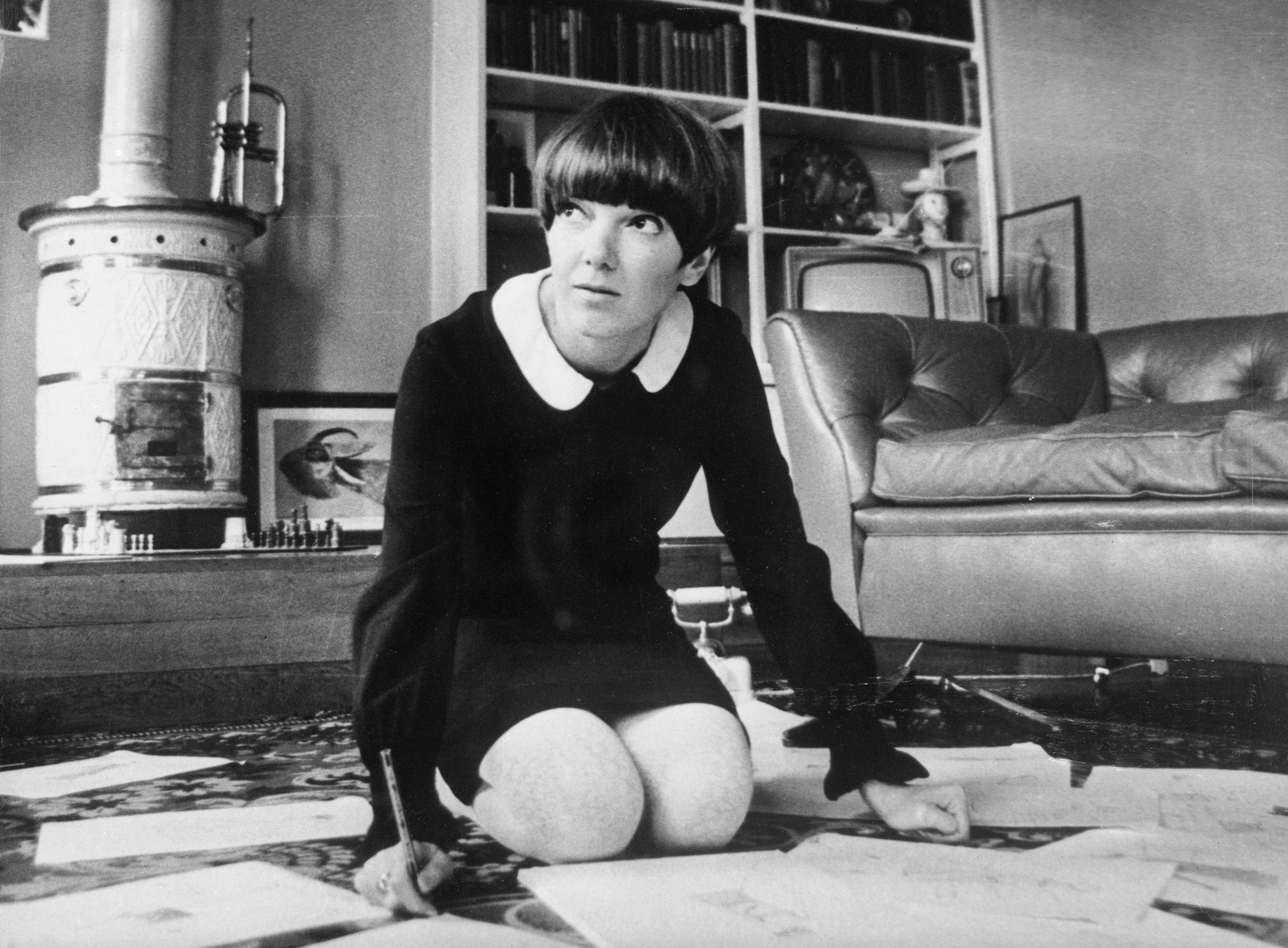 A black and white photo of a woman in a short dress with a bob haircut kneeling on her floor.