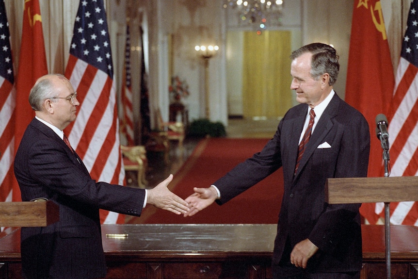 The two men, in suits, shake hands in front of a stage set with US and Soviet flags