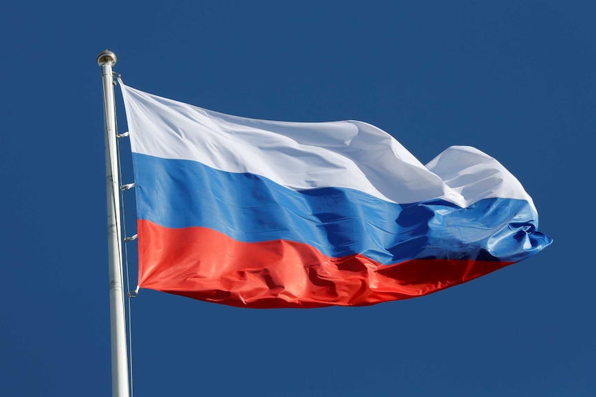 The Russian flag flies on a pole in front of a blue sky.