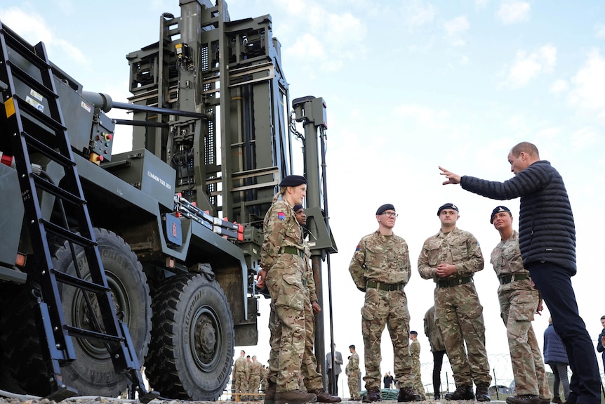 William gestures as he speaks to a group of uniformed soldiers around him. To the right is a large military vehicle