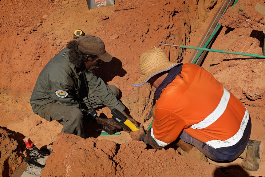 A ranger and a builder crouch down in the dirt to work on the construction of a crocodile farm.