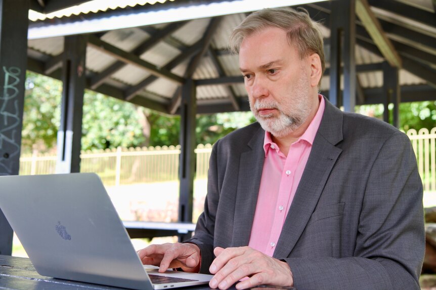 Paul Sadler wearing a grey jacket, using a laptop at an outdoor table.