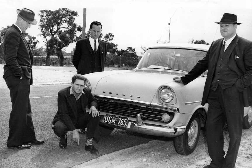 Black and white pic of Eric Edgar Cooke, crouching in front of a police car with detectives.