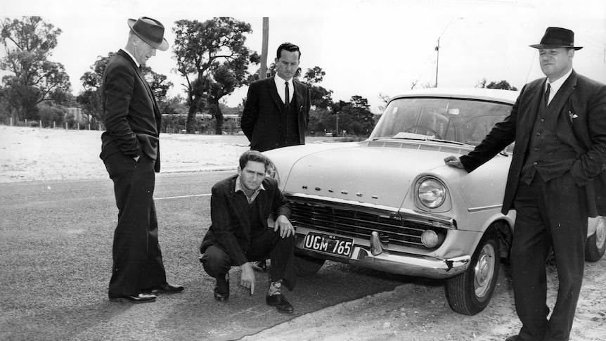 Black and white pic of Eric Edgar Cooke, crouching in front of a police car with detectives.