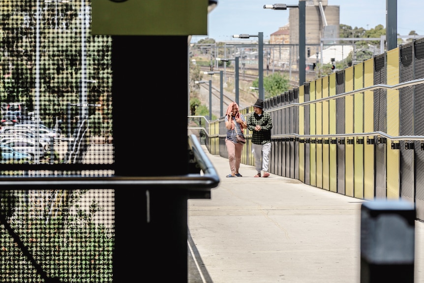 Two people chat as they walk up a pedestrian path. There are fences on either side of the path and there is no cover