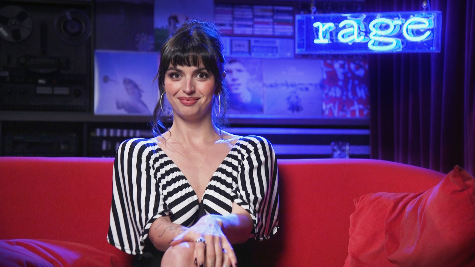 Rebecca Black sits smiling on a red couch in front of a neon blue sign that reads rage