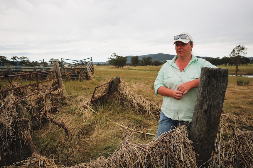 A woman leans against a wooden post in front of vegetation wrapped around a leaning fence.