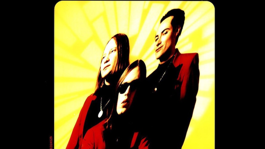 Three men in red suits stand against a yellow background and the image has been warped to tilt to one side