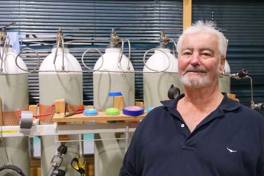 A man with white hair and a white beard faces the camera, with four gas cylinders in the background.