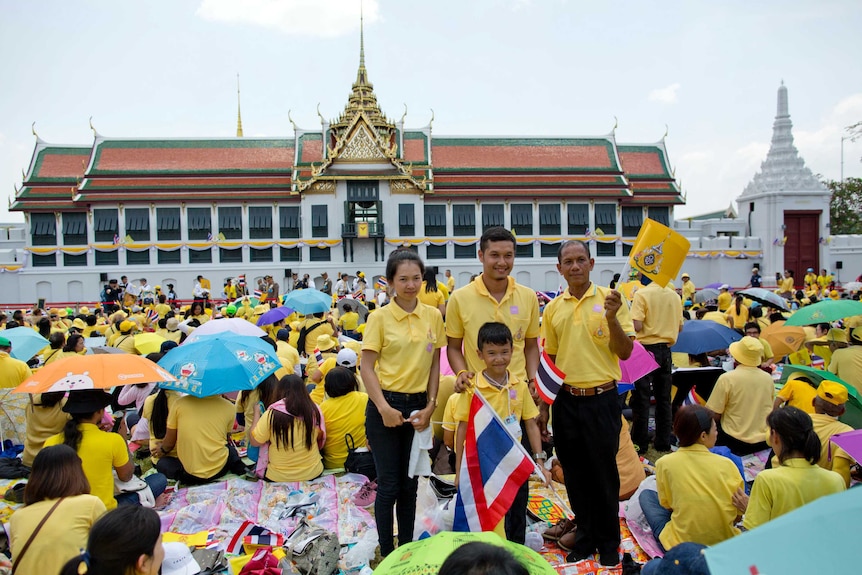 You see throngs of people dressed in yellow filling a square in front of an ornate Thai Royal building.