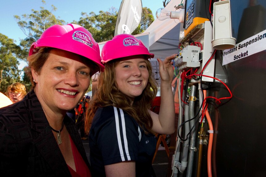Two women in bright pink hard hats and reflective vests standing next to an electrical switch board.