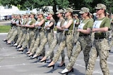 A group of women in army fatigues and high-heel shoes march together on a sunny day.