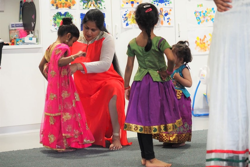 An Indian dance teacher helps a young girl adjust her pink and gold sari as three other young girls watch on.