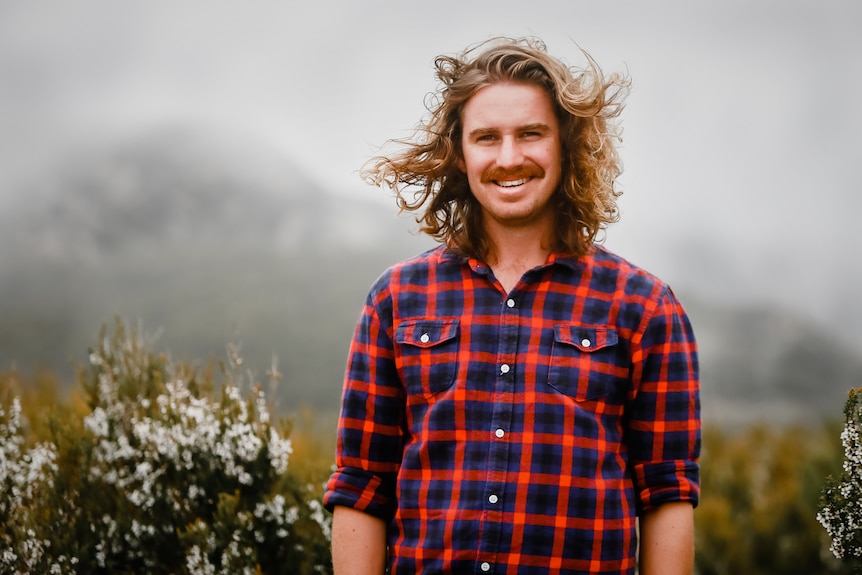 Man wearing bright checked shirt with long hair blowing in wind smiling to camera while standing in shrubby vegetation.