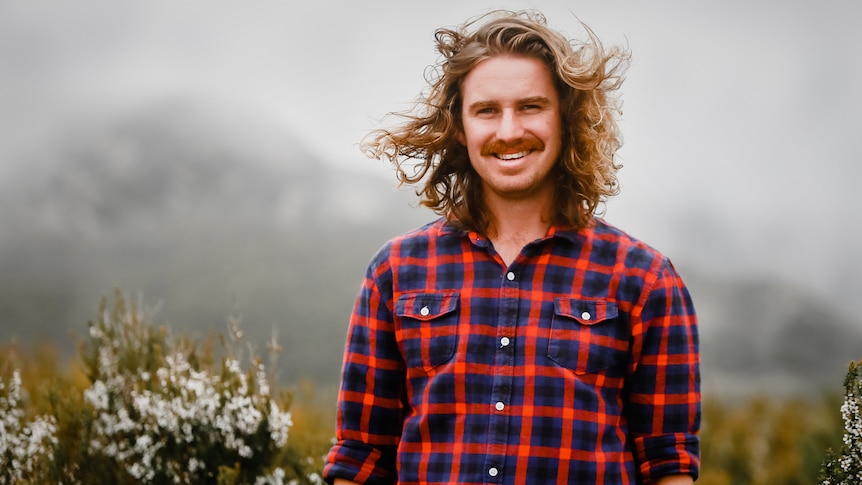 Man wearing bright checked shirt with long hair blowing in wind smiling to camera while standing in shrubby vegetation.