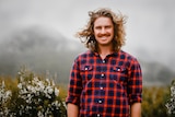 Man wearing bright checked shirt with long hair blowing in wind smiling to camera while standing in shrubby vegetation.