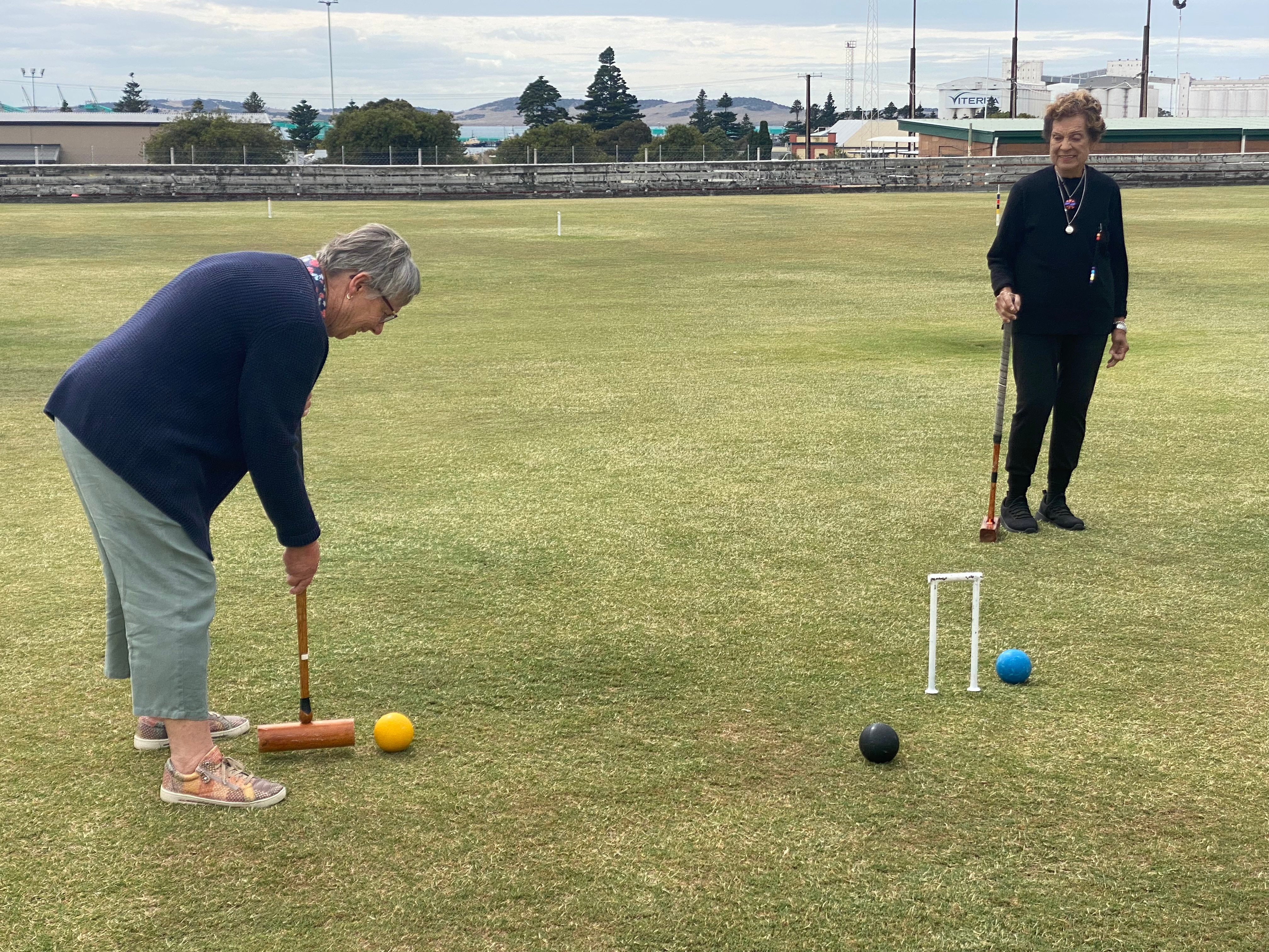 Uma mulher jogando croquet com uma bola amarela, mirando em um aro. Outra mulher assistindo.