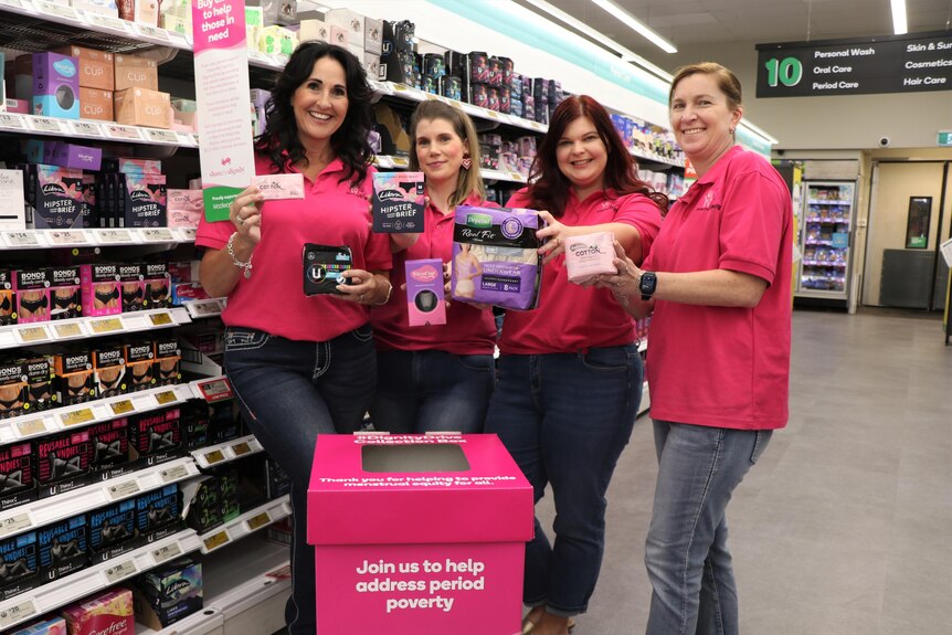 four women wearing pink shirts hold sanitary products in a super market aisle.