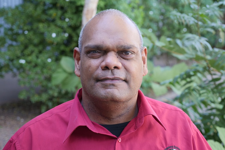 A Aboriginal man with close-shaved grey hair standing in front of a leafy garden