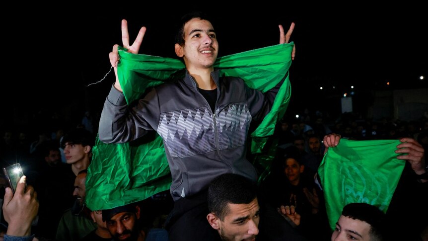 A young boy with a green flag makes the peace sign while sitting on a man's shoulders