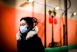 A Chinese woman in a fur-trimmed coat and blue face mask walks down a street near an orange wall