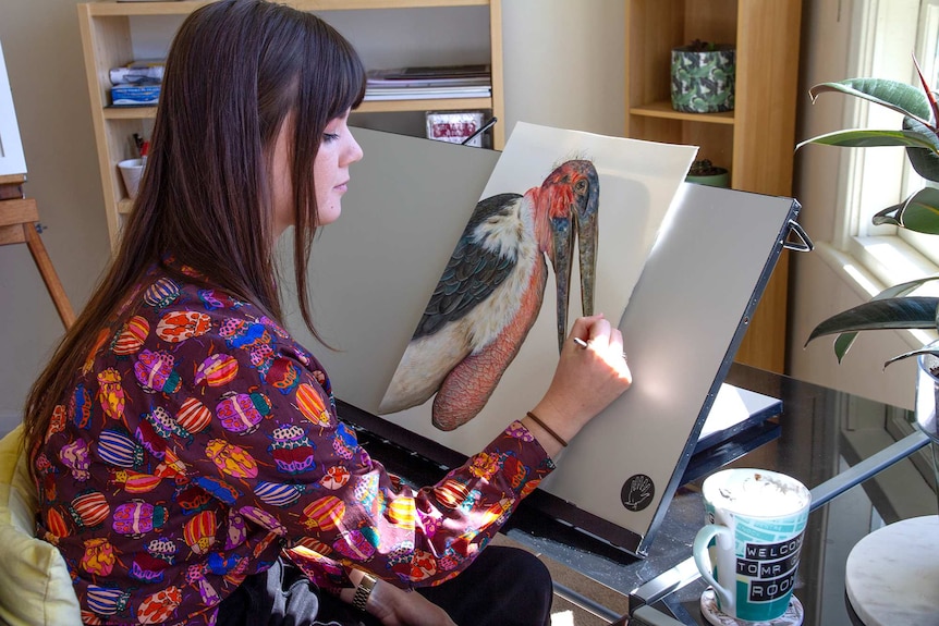 A young woman works by hand on an illustration of an exotic looking bird.