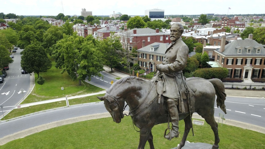 A statue of Robert E Lee on a horse, with red-brick houses and tree-lined streets in the background.