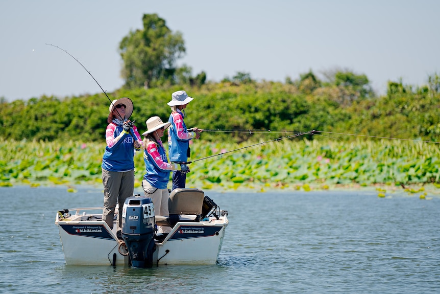 Three women stand on a boat with fishing lines in the water.