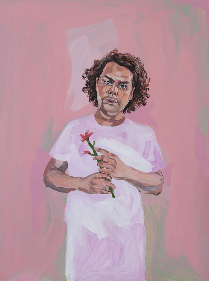 Thompson stands against a pink background, wearing a t-shirt in a lighter shade of pink, holding a small flower.