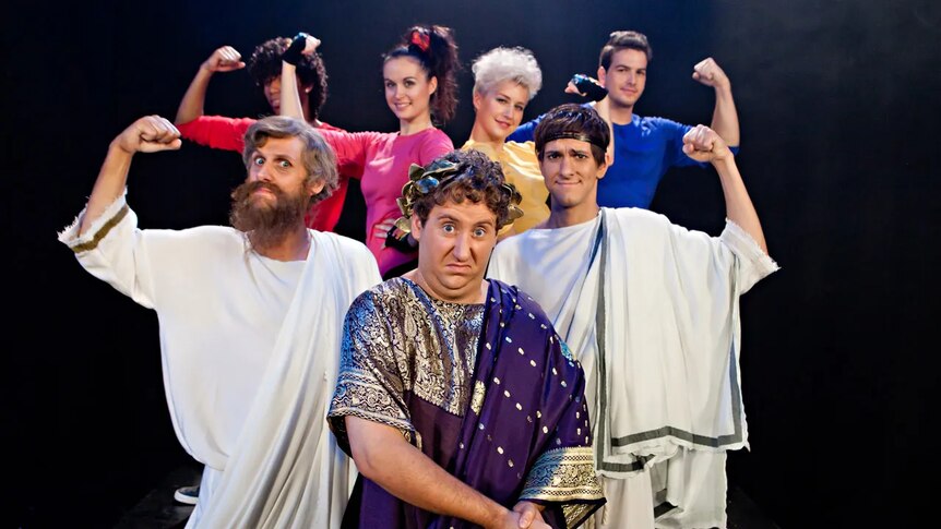 A man is dressed as Julius Caesar and six people in togas all raise their arms in a bicep curl