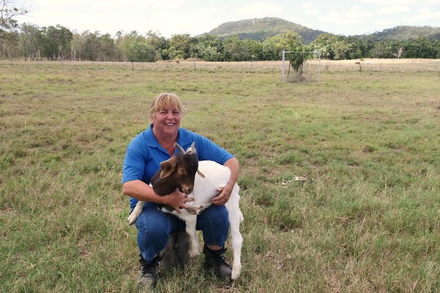 woman kneels holding a goat in a paddock