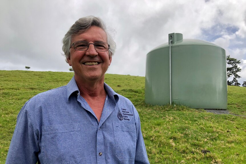 A man smiles at the camera in a paddock with a water tank behind him.