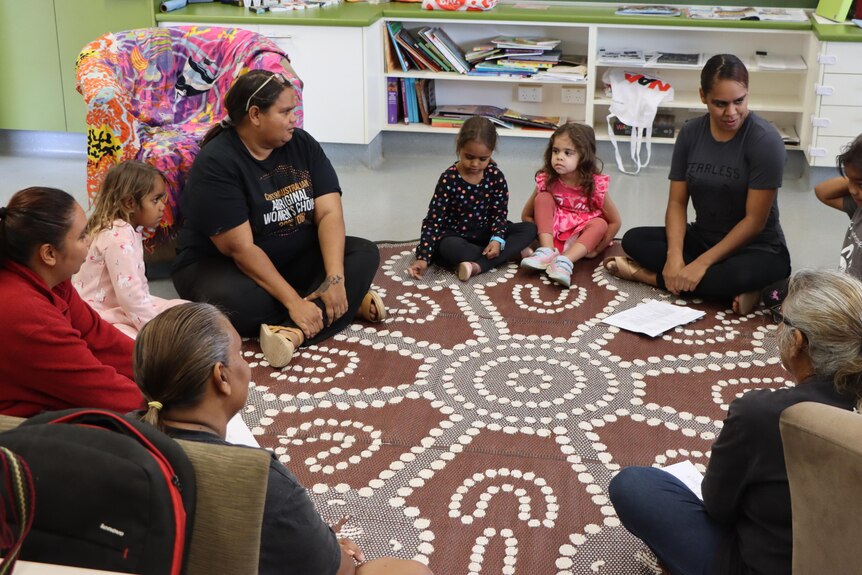 A group of kids and women sit in a circle on the floor 