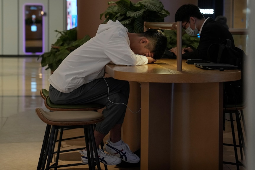 A young man wearing jeans and a white hoodie rests his head on a table, while across from him someone works.