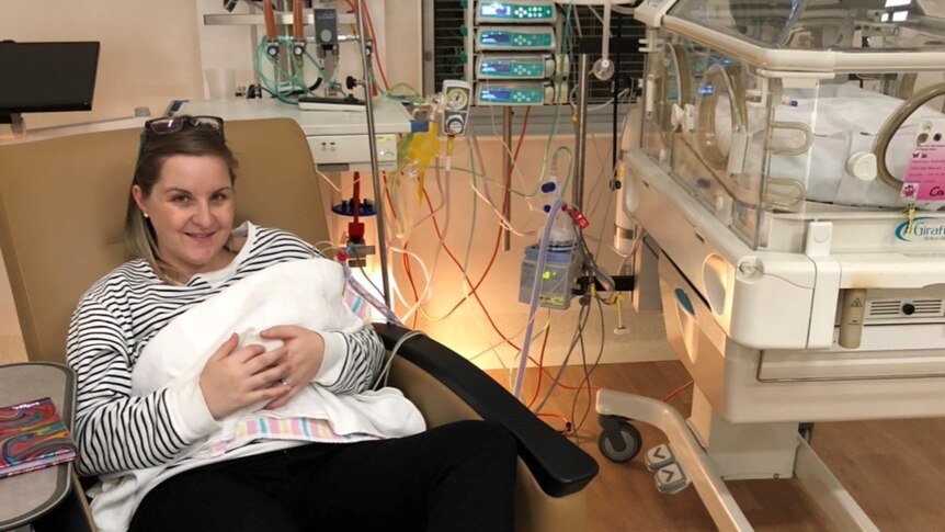 Mum sits in a chair and holds her baby in an intensive care unit with a humidicrib.