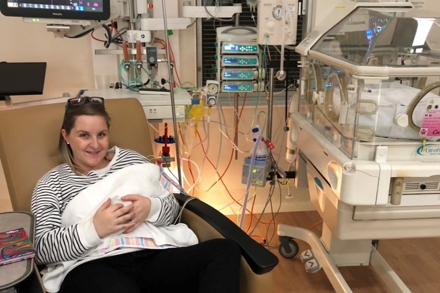 Mum sits in a chair and holds her baby in an intensive care unit with a humidicrib.