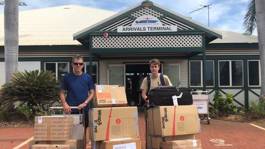 Father and son standing with a number of carboard boxes outside the arrivals terminal at Broome airport