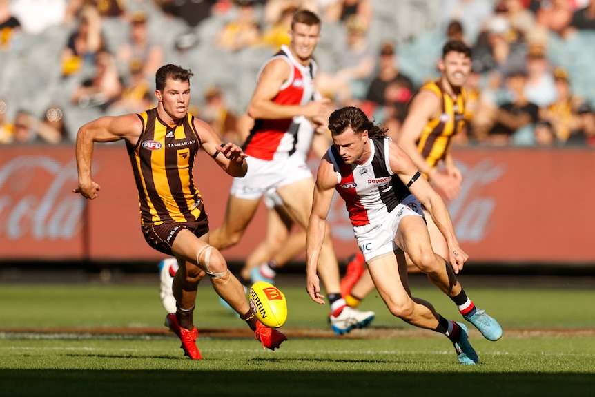 A St Kilda player bends down to grab a bouncing ball as a Hawthorn midfielder closes in on him.
