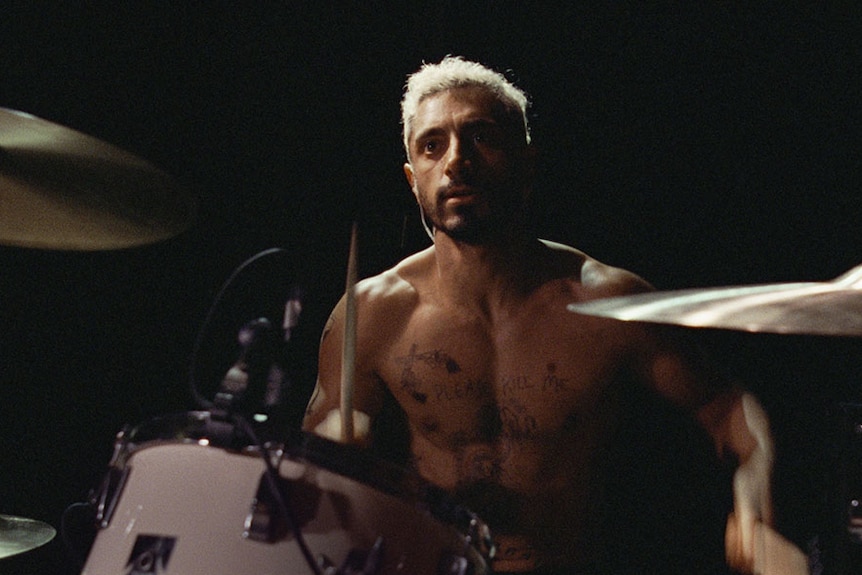 Man bare-chested with tattoos sitting at drum kit playing.