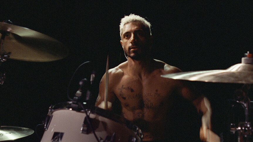 Man bare-chested with tattoos sitting at drum kit playing.