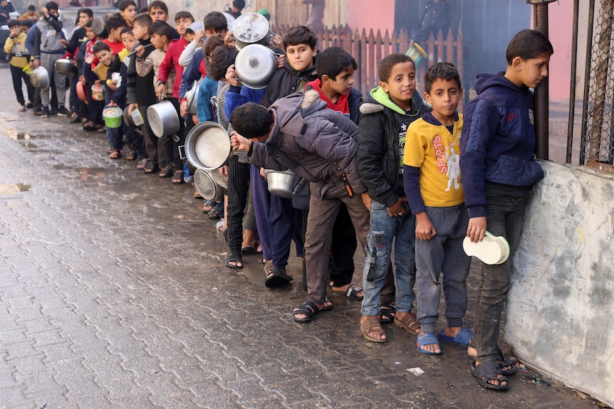 Palestinian children carry pots as they queue to receive food on a damp street.