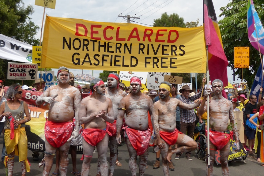 Aboriginal men in traditional paint and clothes march under a sign reading Declared Northern Rivers Gasfield Free.