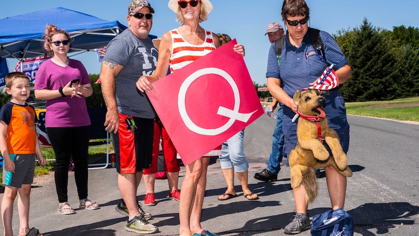 Five people stand outdoors. A woman in the centre carries a red sign that says "Q".