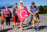 Five people stand outdoors. A woman in the centre carries a red sign that says "Q".