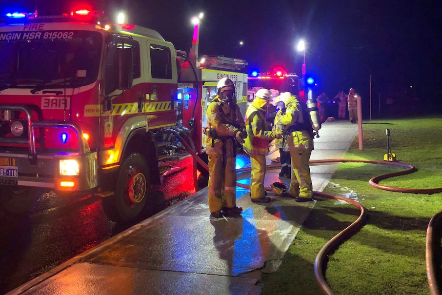 Firefighters in front of fire trucks at night, with hoses on the ground.